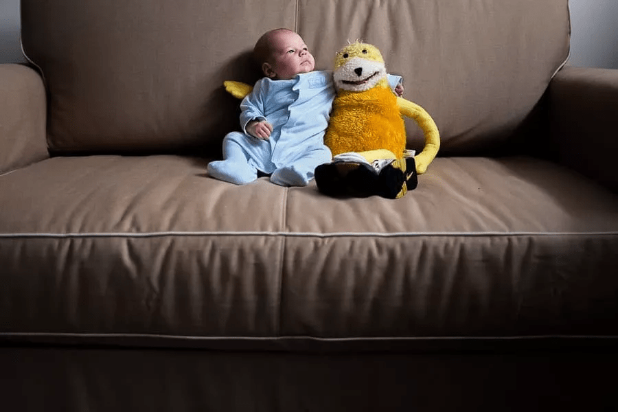 یک نوزاد که روی مبل نشسته و عروسکش را بغل کرده است